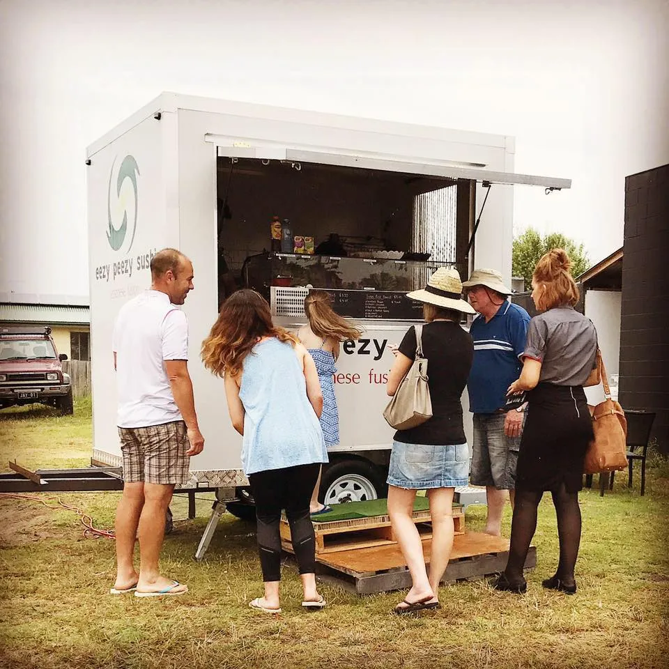 A group of customers gathered at the Eezy Peezy Sushi food truck at an outdoor market, with the truck branding visible and people ordering from the window.